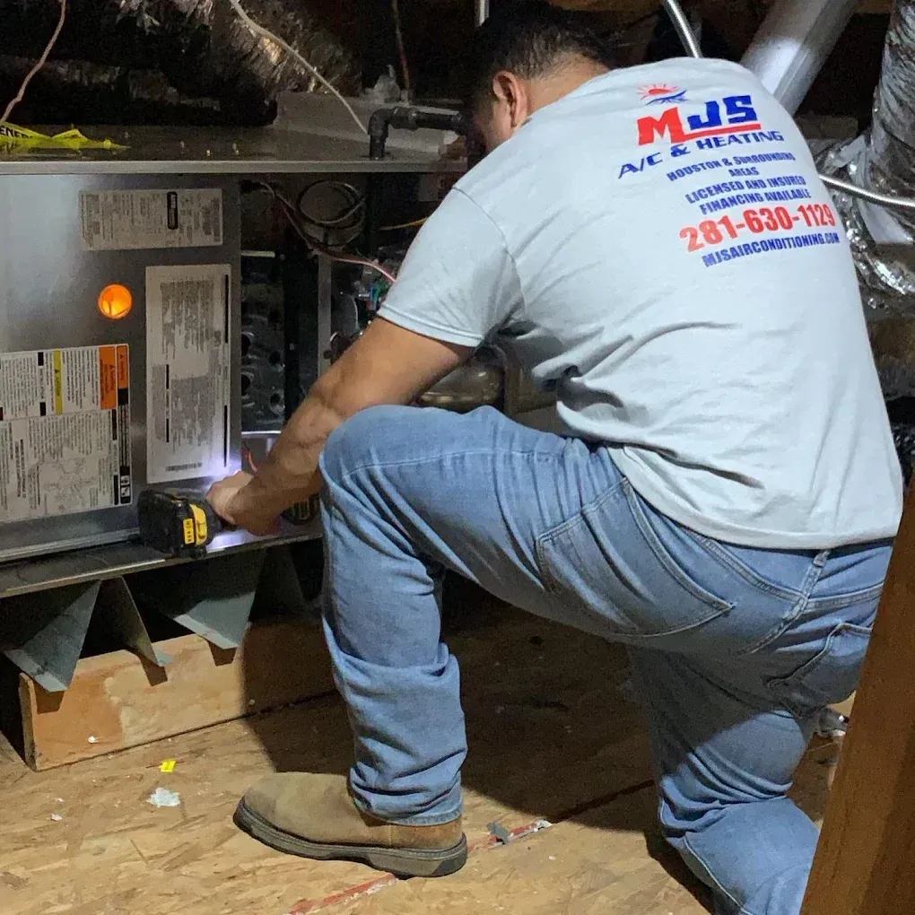 HVAC technician kneeling to work on a unit in an attic, wearing a company shirt with a phone number.