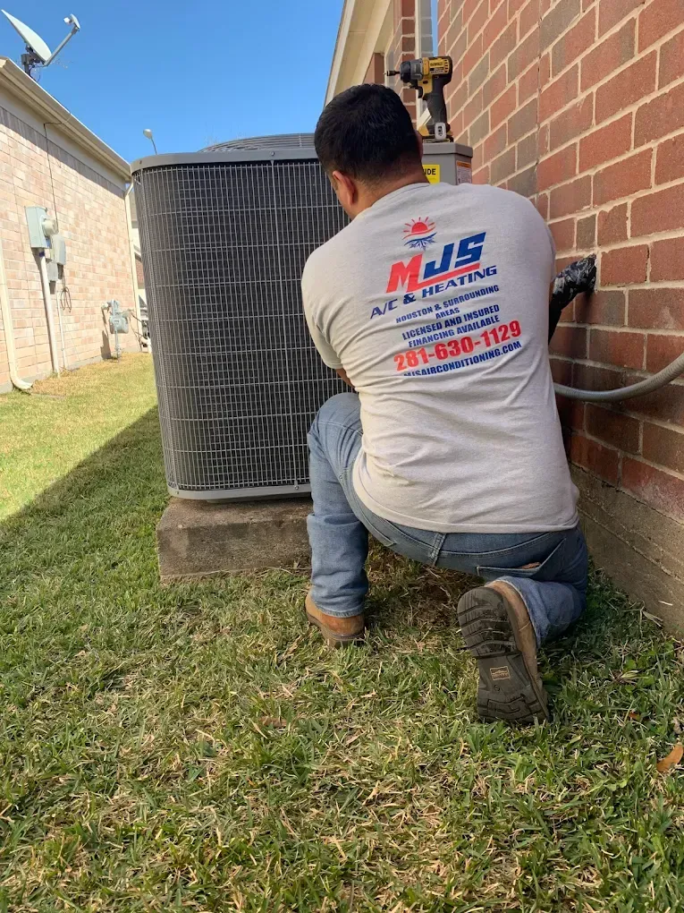 HVAC technician kneeling near an AC unit and brick wall. Green grass. Gray shirt, jeans, and work boots.