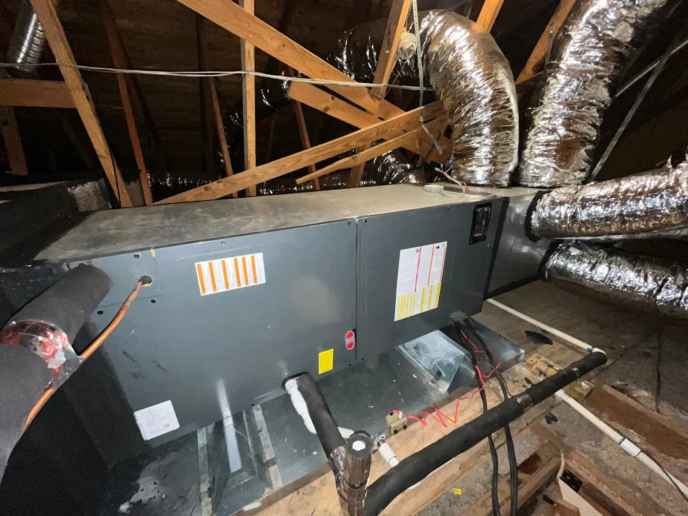 HVAC unit in an attic, with ducts and wooden supports. Gray metal box with vents and insulation.