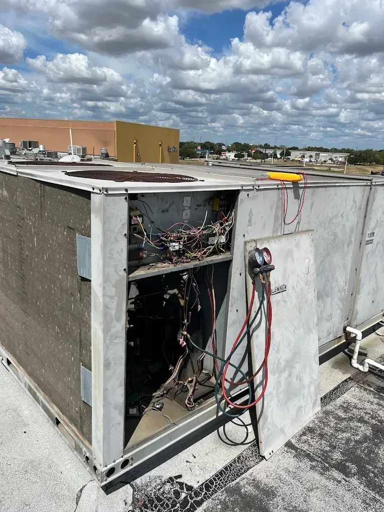 HVAC unit on a rooftop with open access panel, wires, and gauges. Cloudy sky in background.