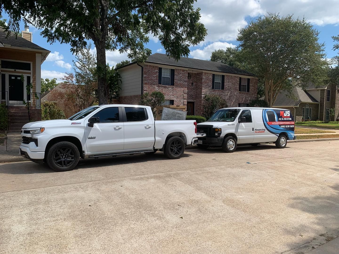 White truck and van parked on street in front of brick buildings, under trees and a partly cloudy sky.