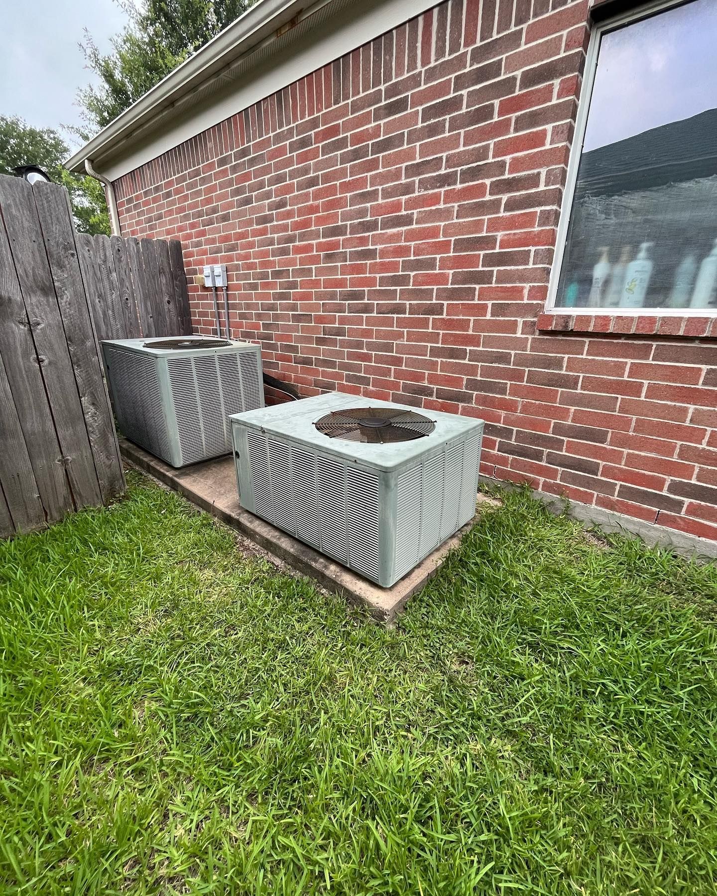 Two air conditioning units on concrete pads beside a brick building and a wooden fence.