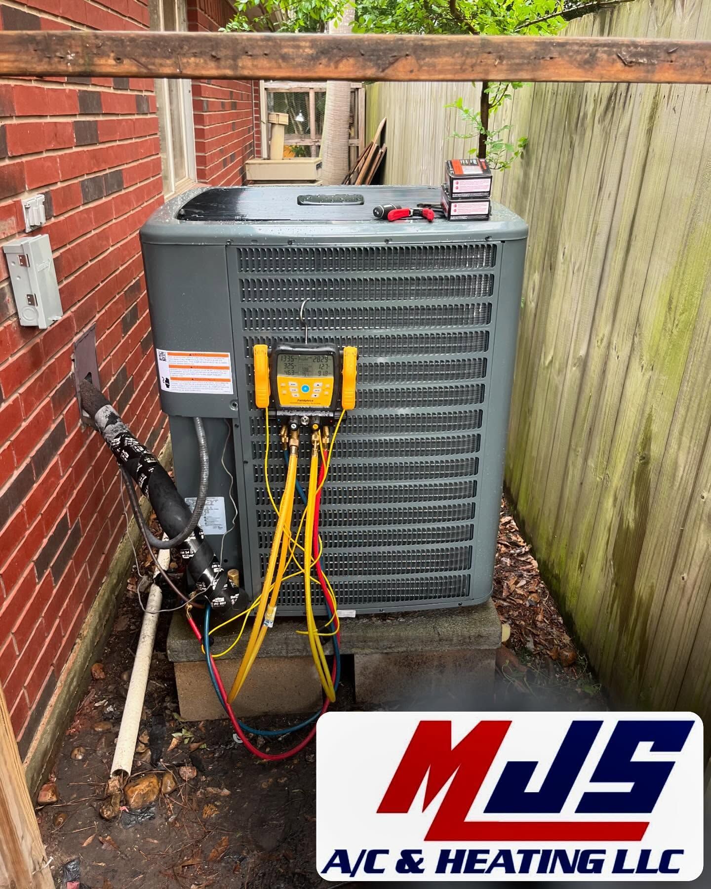 HVAC unit with gauges, wires, and tools. Located near a brick wall and wooden fence. MJS A/C & Heating LLC logo in foreground.