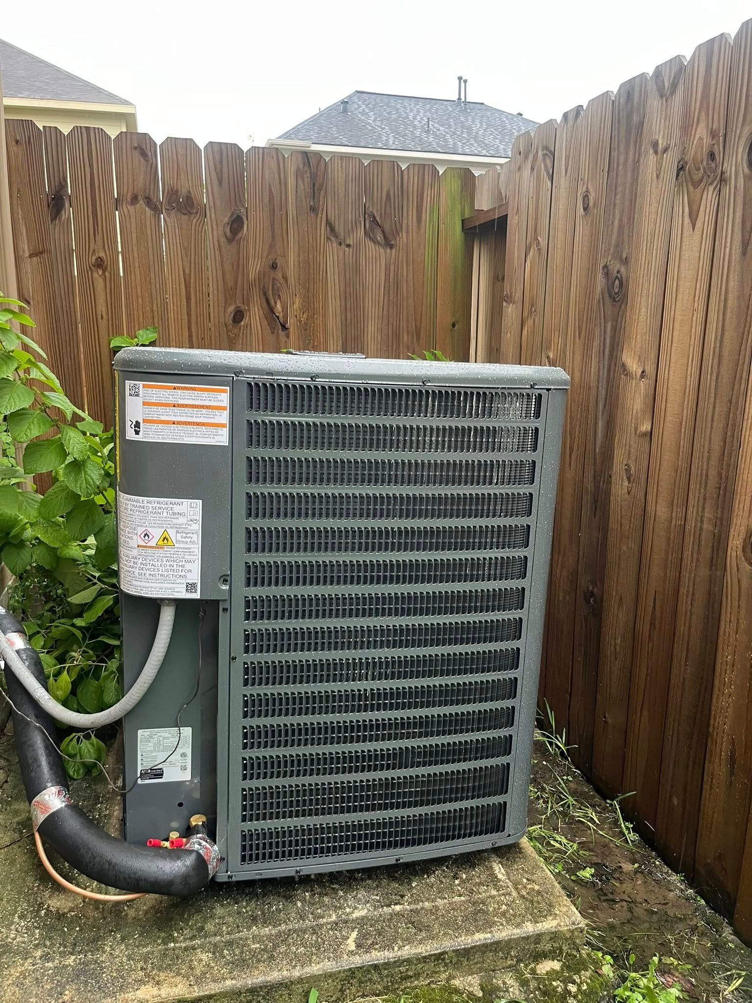 Air conditioning unit next to a wooden fence. Gray metal, black hose, green vines.