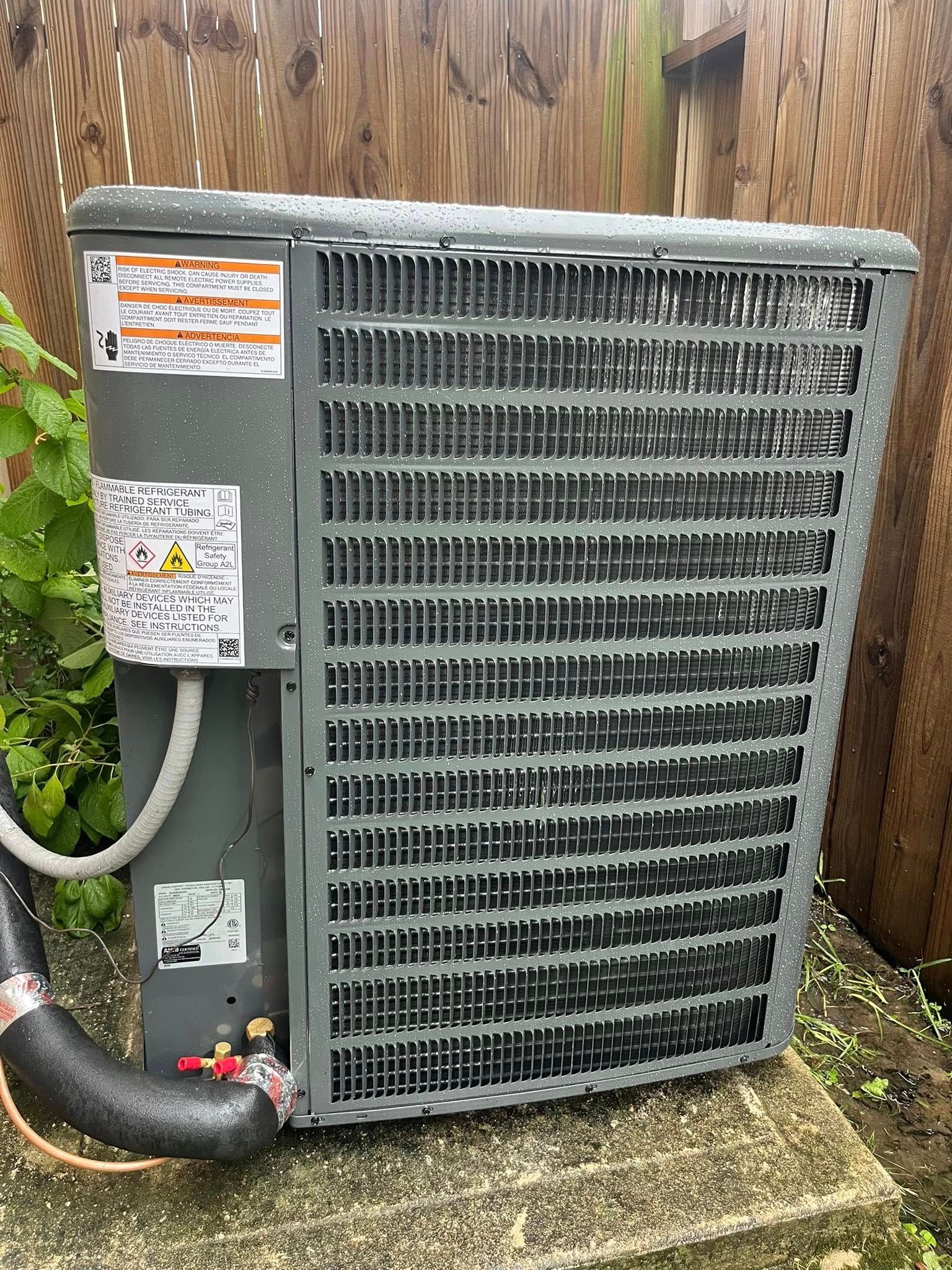 Outdoor air conditioning unit against a wooden fence. Gray metal with a black hose and red valve.
