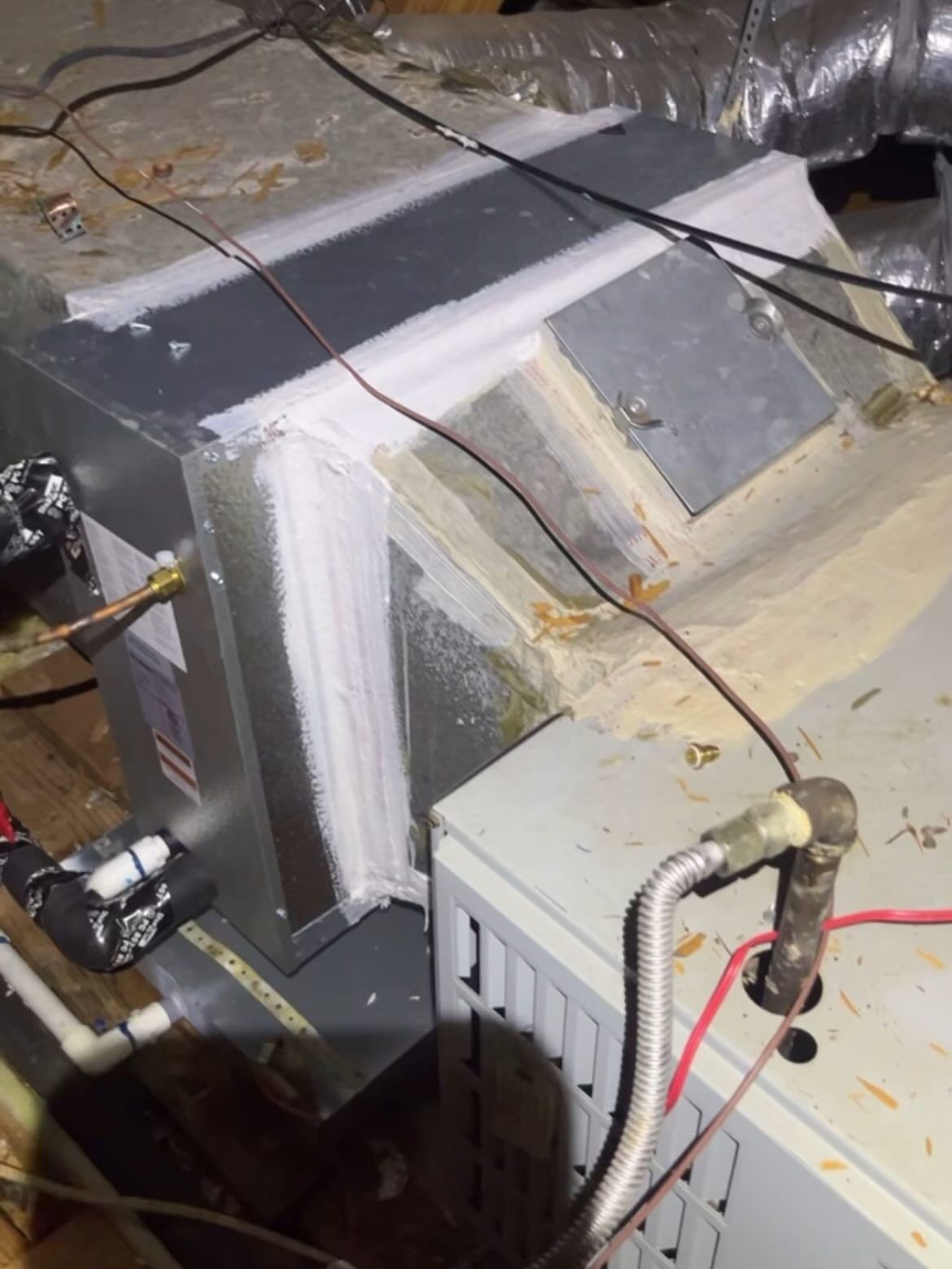 HVAC unit components in an attic, with metallic ductwork and insulation. Wires and pipes visible.