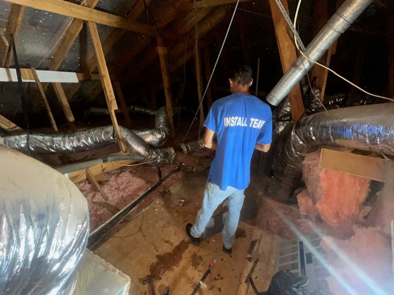 Person in a blue shirt in an attic, inspecting ductwork. Setting is dimly lit, with insulation and exposed beams visible.
