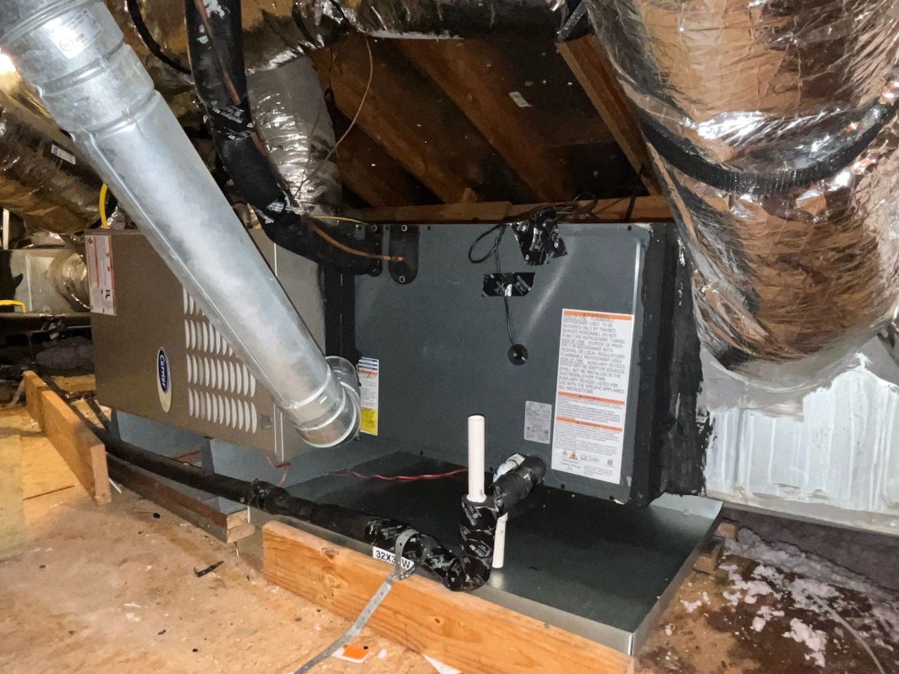 HVAC system in an attic. Gray metal unit with ductwork and insulation, set on wooden beams.