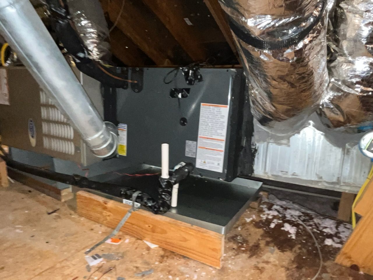 HVAC unit in an attic with ductwork and piping. The unit is grey and resting on wooden supports.