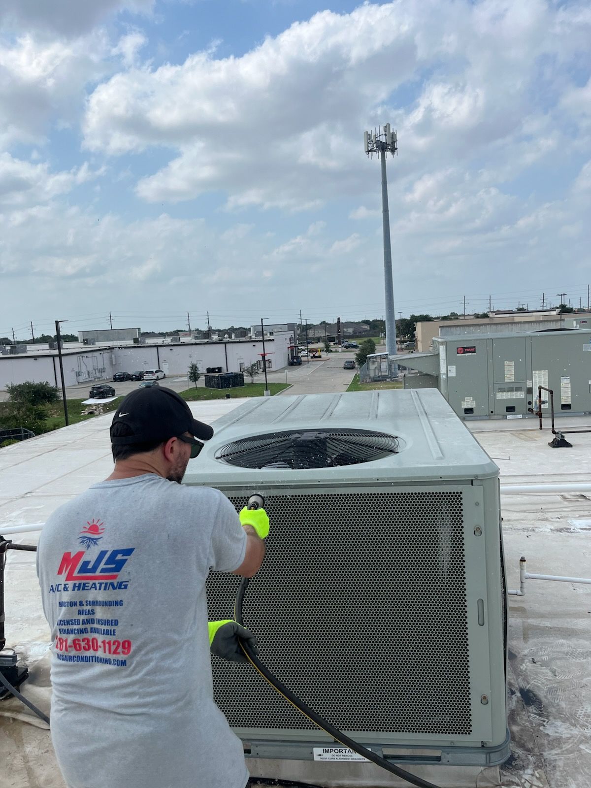HVAC technician spraying an air conditioner on a rooftop. Bright, cloudy sky.