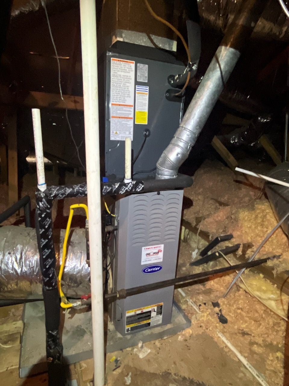 Furnace in an attic. Gray metal unit with ductwork, electrical wiring, and insulated pipes visible.