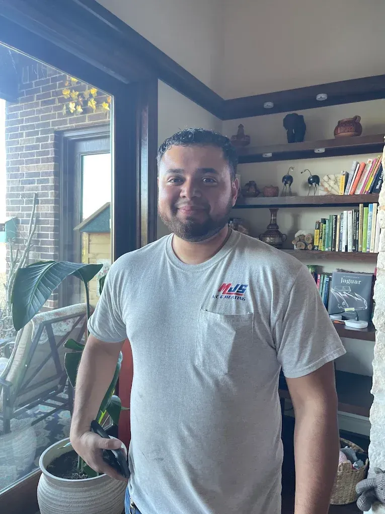 Man in gray t-shirt smiles near window, with a bookshelf and outdoor view visible.