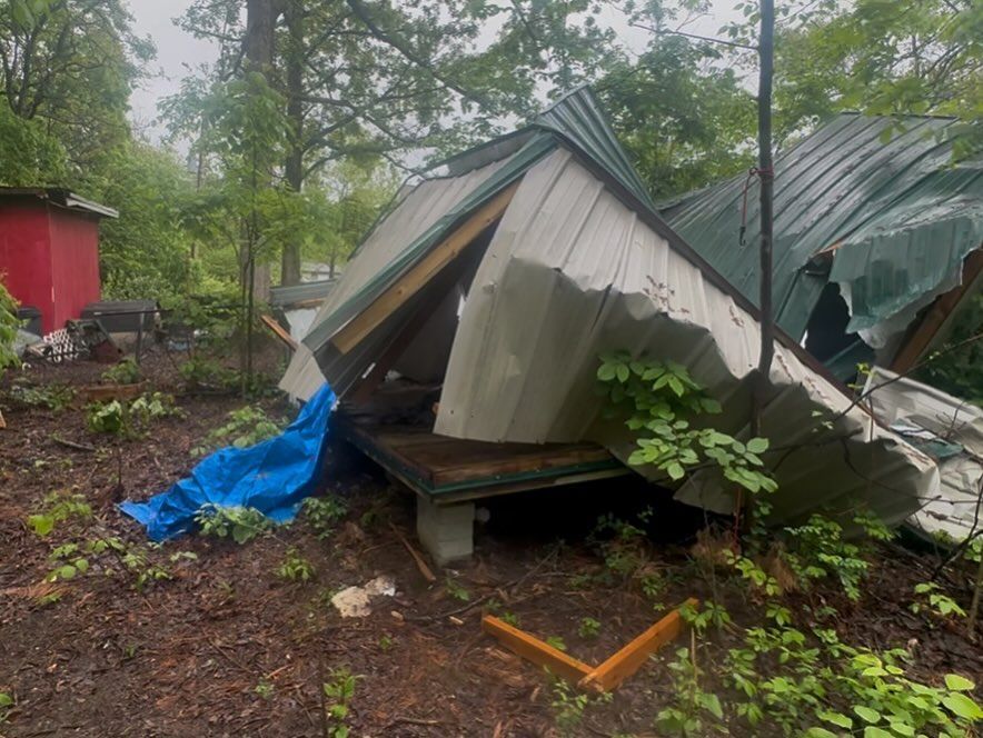 Collapsed metal-roofed shelter amid muddy debris and trees, with a blue tarp nearby.