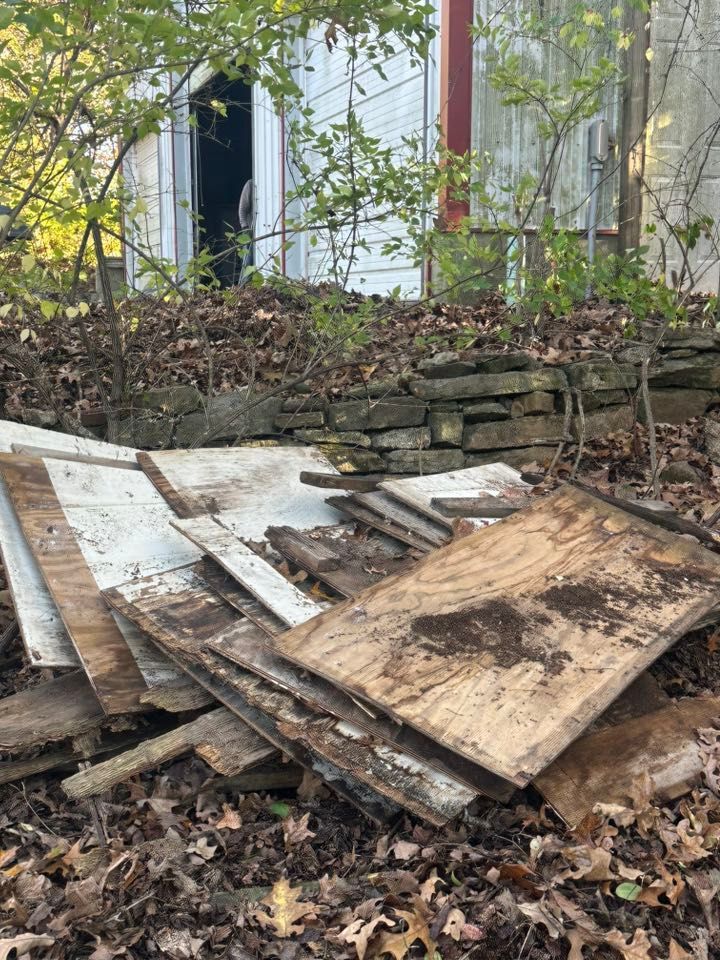 Plywood and scrap wood piled in a leaf-covered yard beside a low stone wall and house.