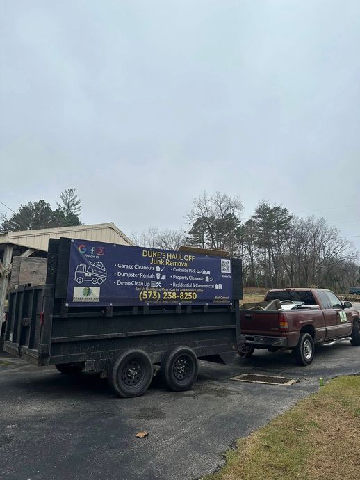 Pickup towing a trailer with a large blue sign along a muddy roadside under cloudy skies