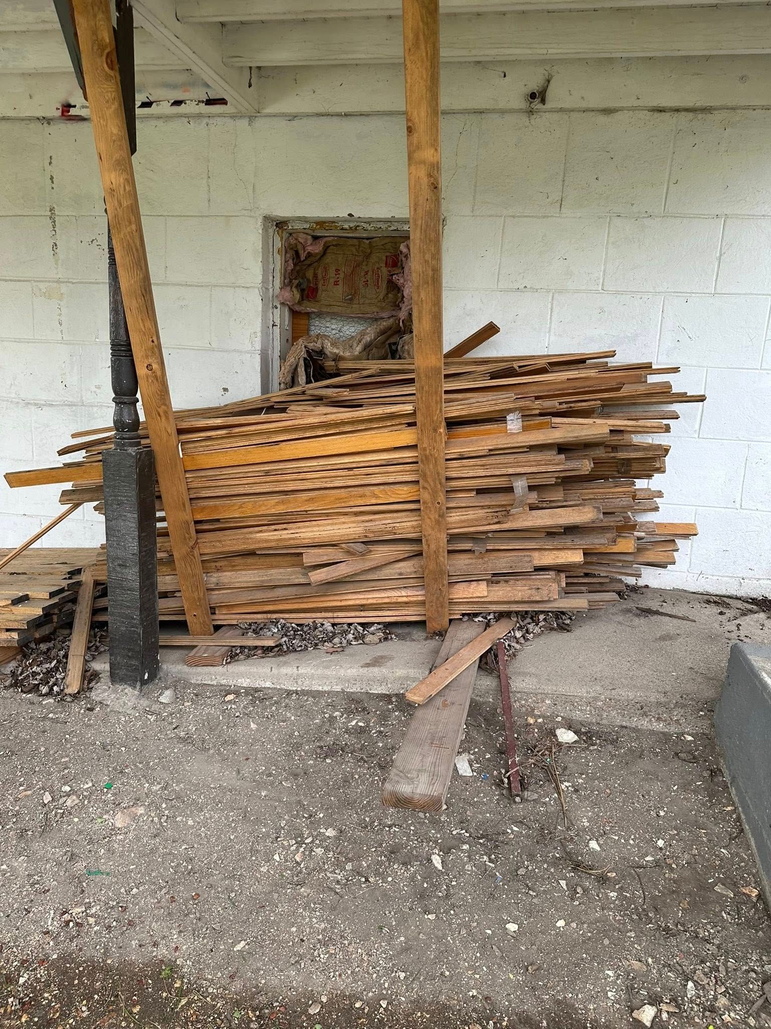 Stack of rough-cut lumber leaning against a wall in a covered outdoor area with wooden posts.