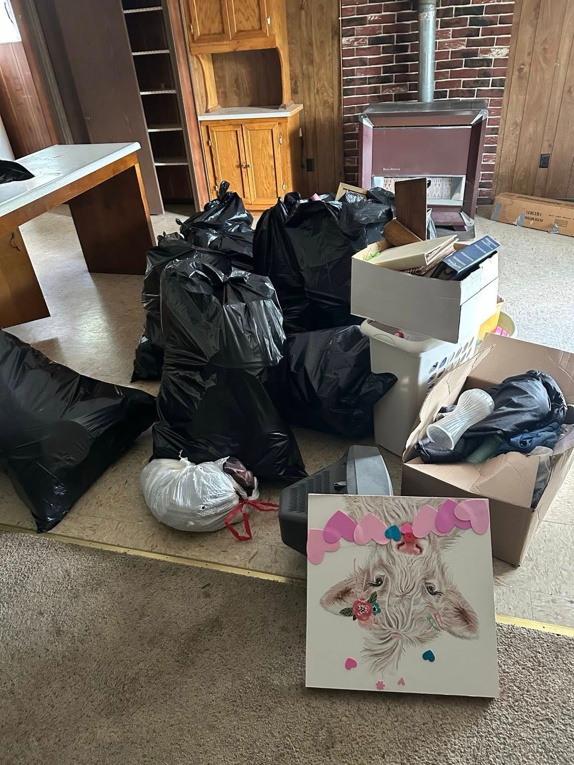 Cluttered room with trash bags, boxes, scattered items, and a floral painting leaning on the floor.