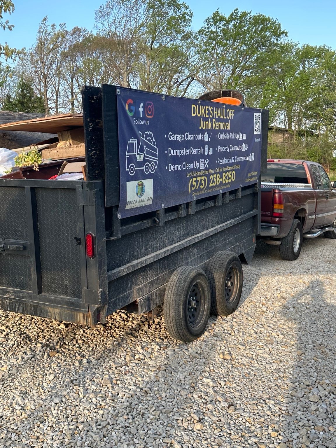 Black dump trailer with landscaping sign parked on gravel beside a pickup truck.