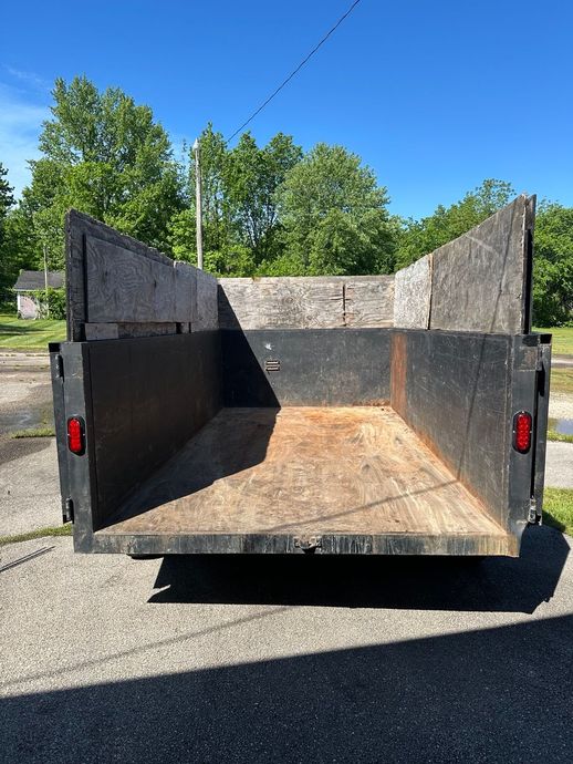 Empty black dump trailer parked outdoors on a sunny street, viewed from the open rear.