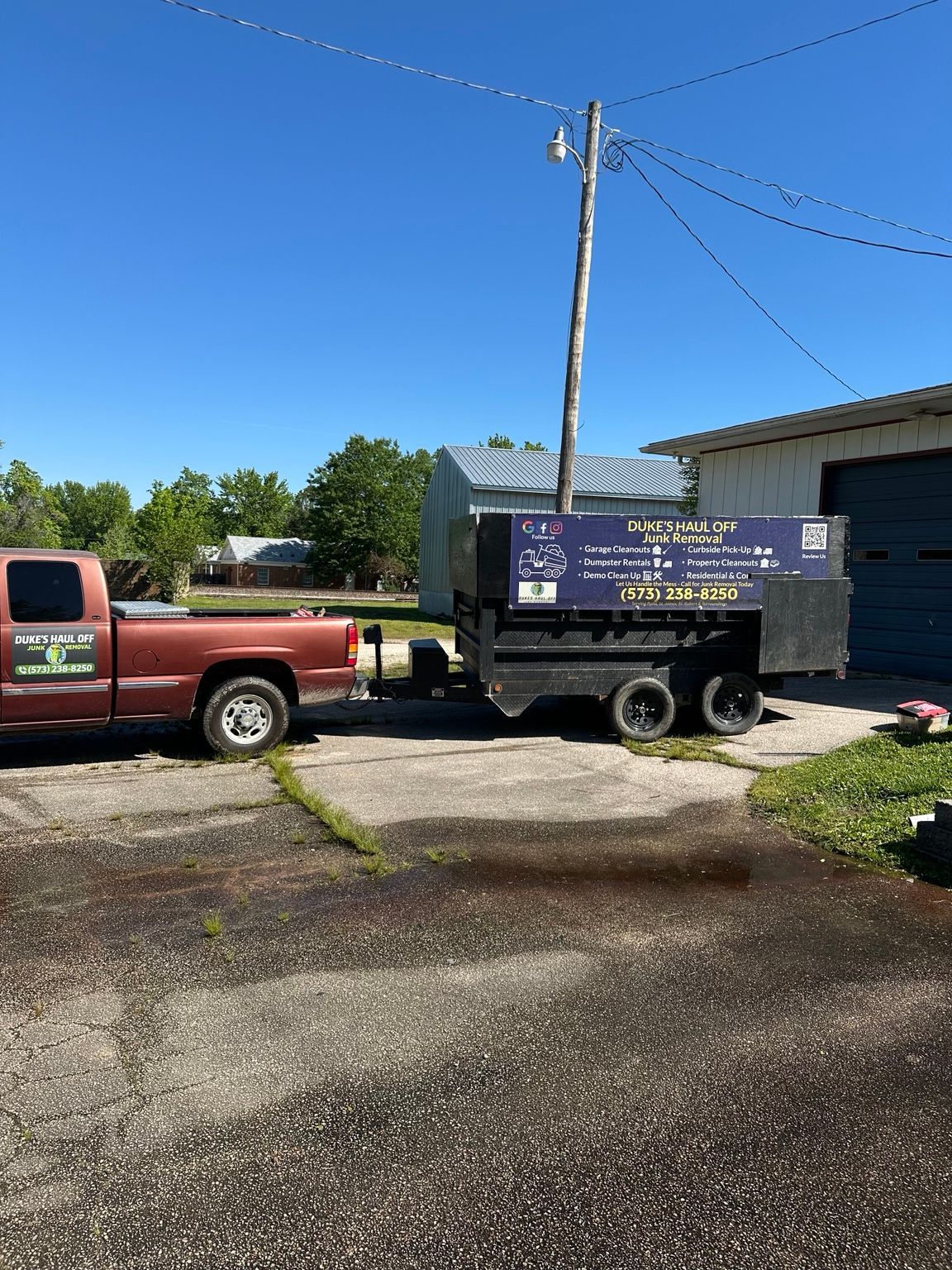 Red pickup truck and black dumpster trailer in a driveway beside a house on a sunny day