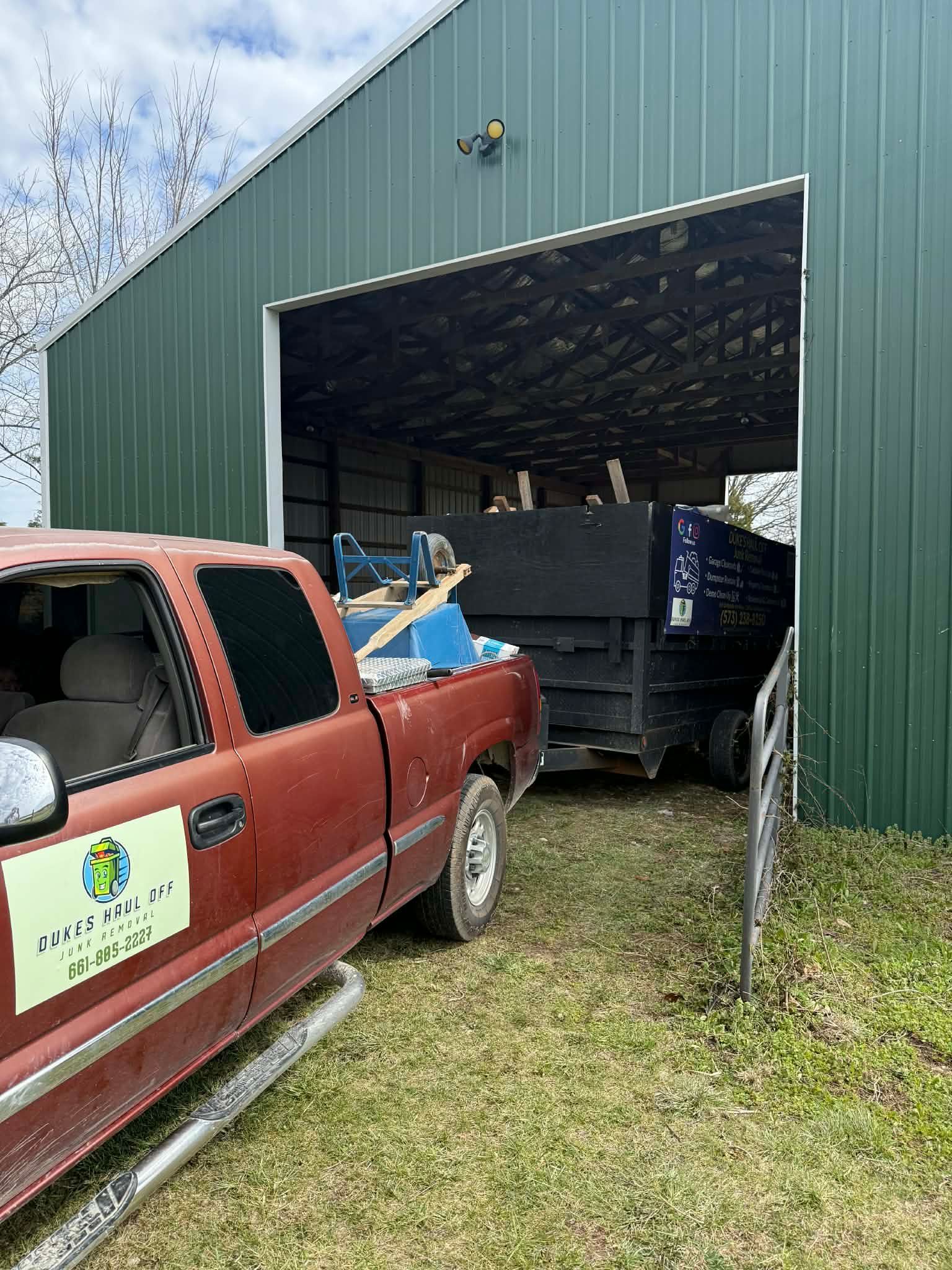 Red pickup truck beside a green metal building with a large open garage door and a dump trailer inside.
