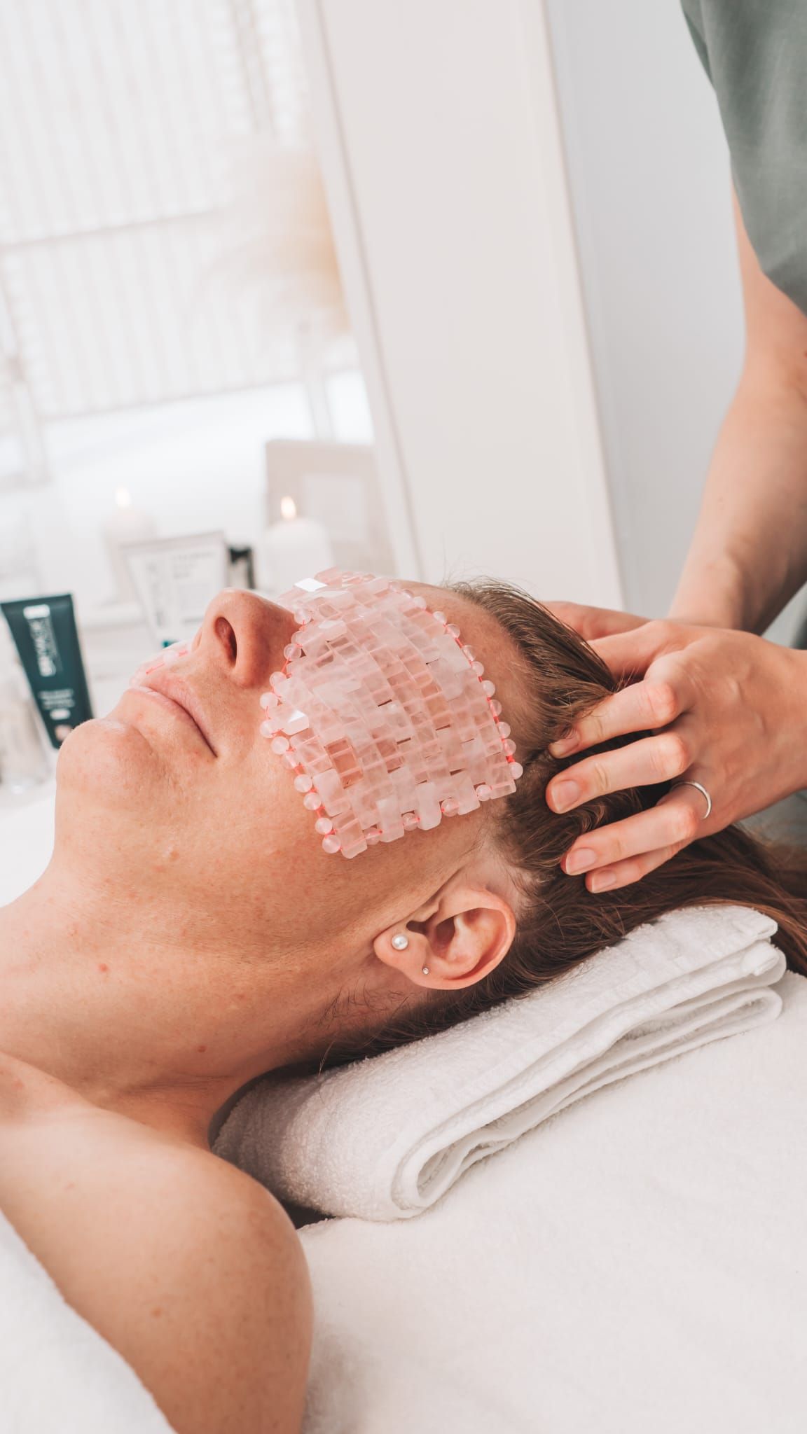 Person receiving a facial treatment; eyes covered with pink crystals, head massage.