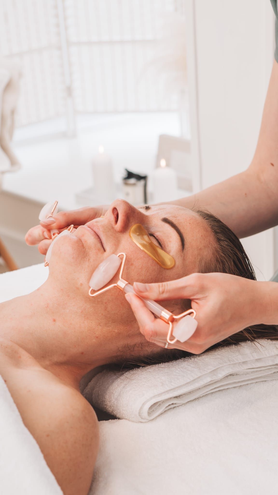 Person receiving facial massage with a rose quartz roller and eye patches. Spa setting.