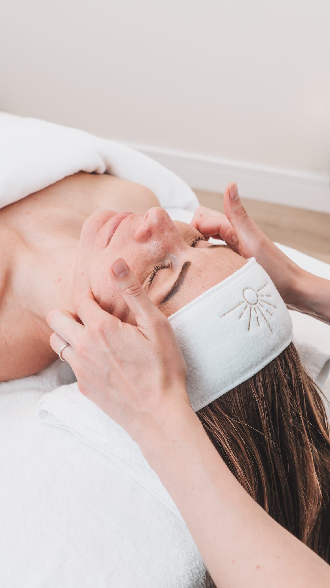 Person receiving a facial massage, lying on a white bed with a headband, eyes closed, hands on face.