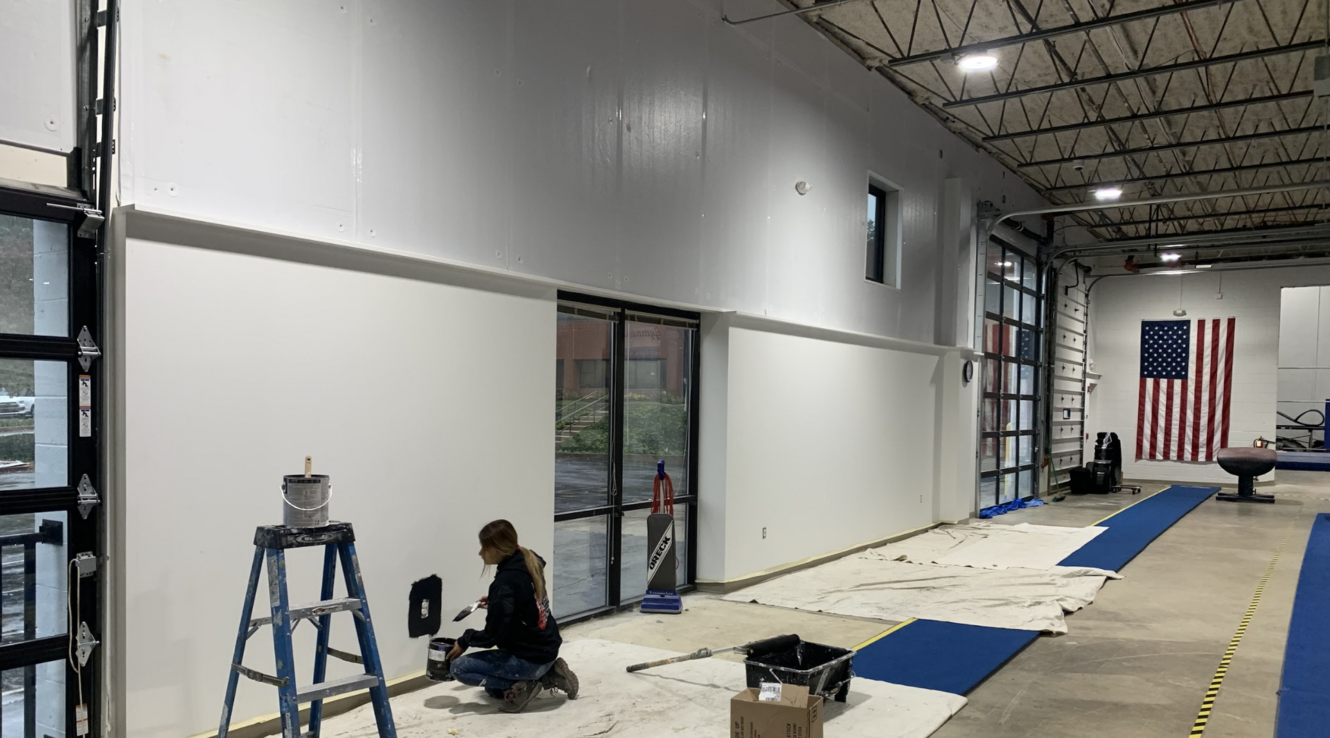 Painter painting the inside of a commercial building. Tarps across the floor and paint bucket on ladder.