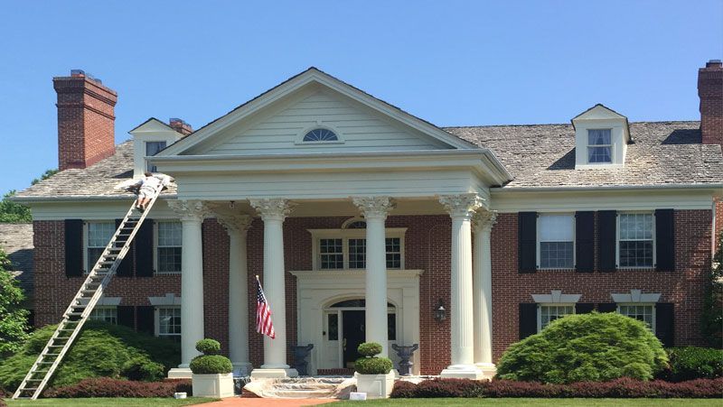 A man is standing on a ladder in front of a large brick house.