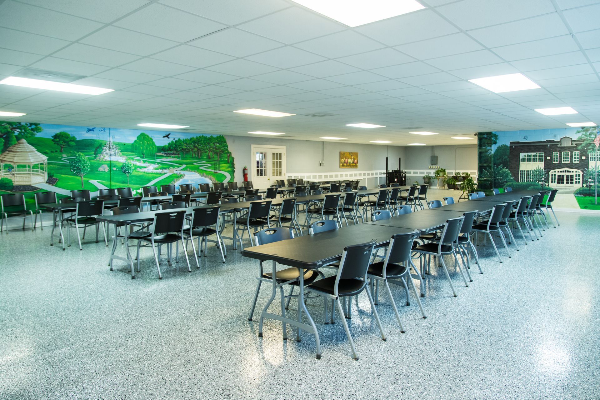 Empty school cafeteria with tables, chairs, mural, and fluorescent ceiling lights