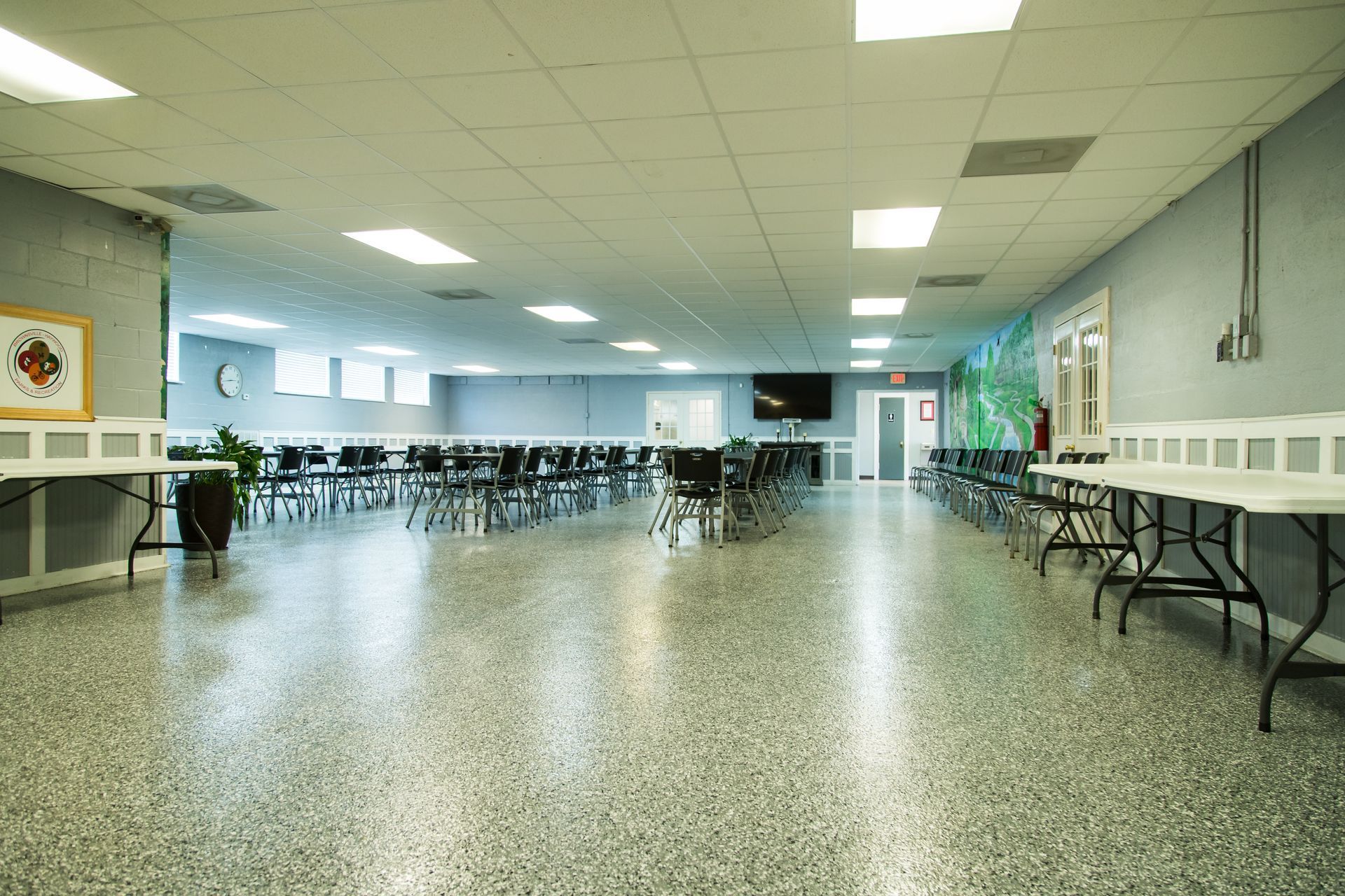 Empty cafeteria or multipurpose room with rows of tables and chairs, gray speckled floor, and bright fluorescent ceiling lights