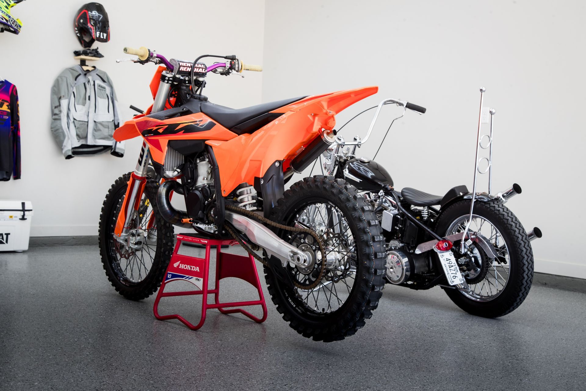 Orange dirt bike and black motorcycle parked in a garage with racing gear on the wall