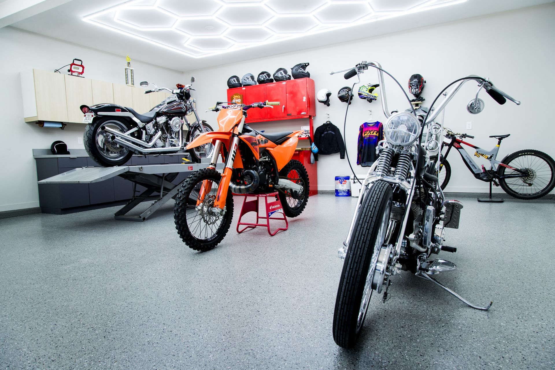 Motorcycles in a bright garage, with an orange dirt bike centered and chrome bikes in the foreground.