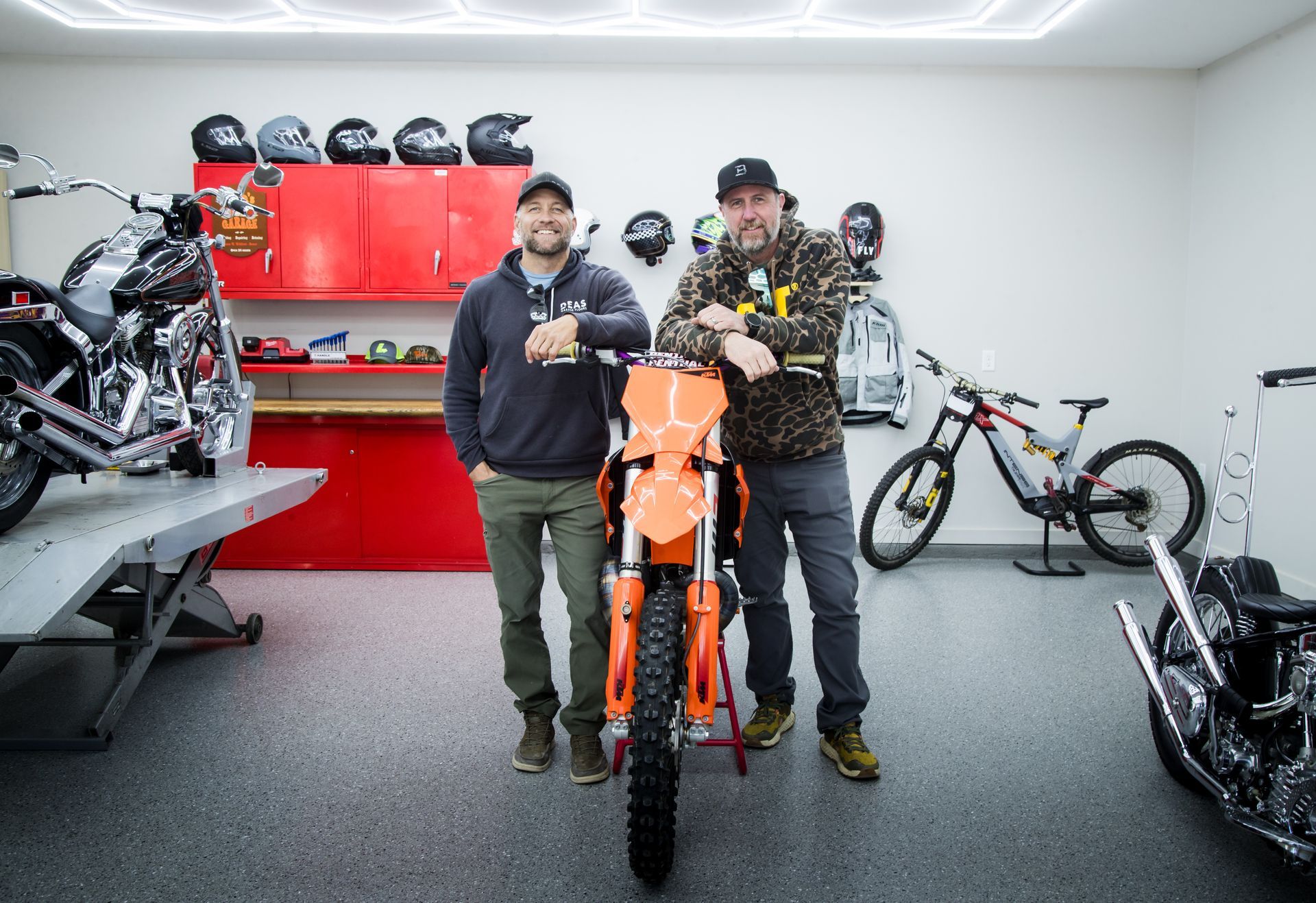 Two men stand with an orange motorcycle in a bright garage, surrounded by bikes and tools.