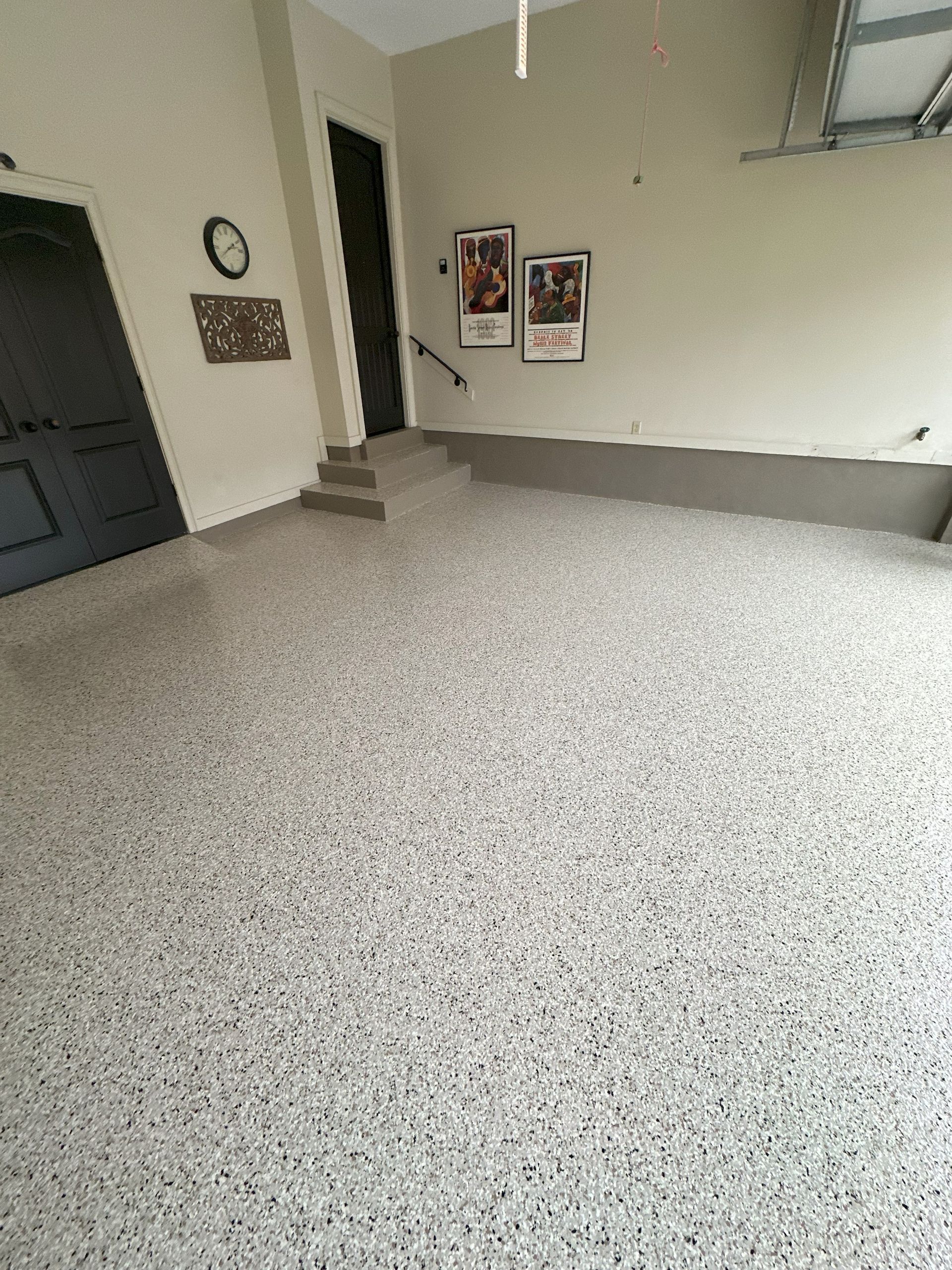 Garage interior with speckled light grey floor, beige walls, black door, and staircase.