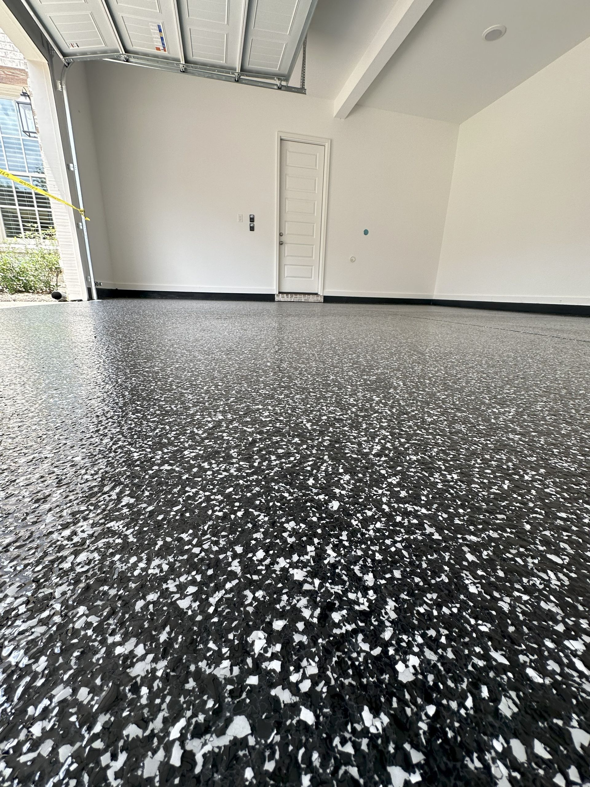 Garage interior with glossy speckled black and white floor. White walls, closed door, and open garage door.