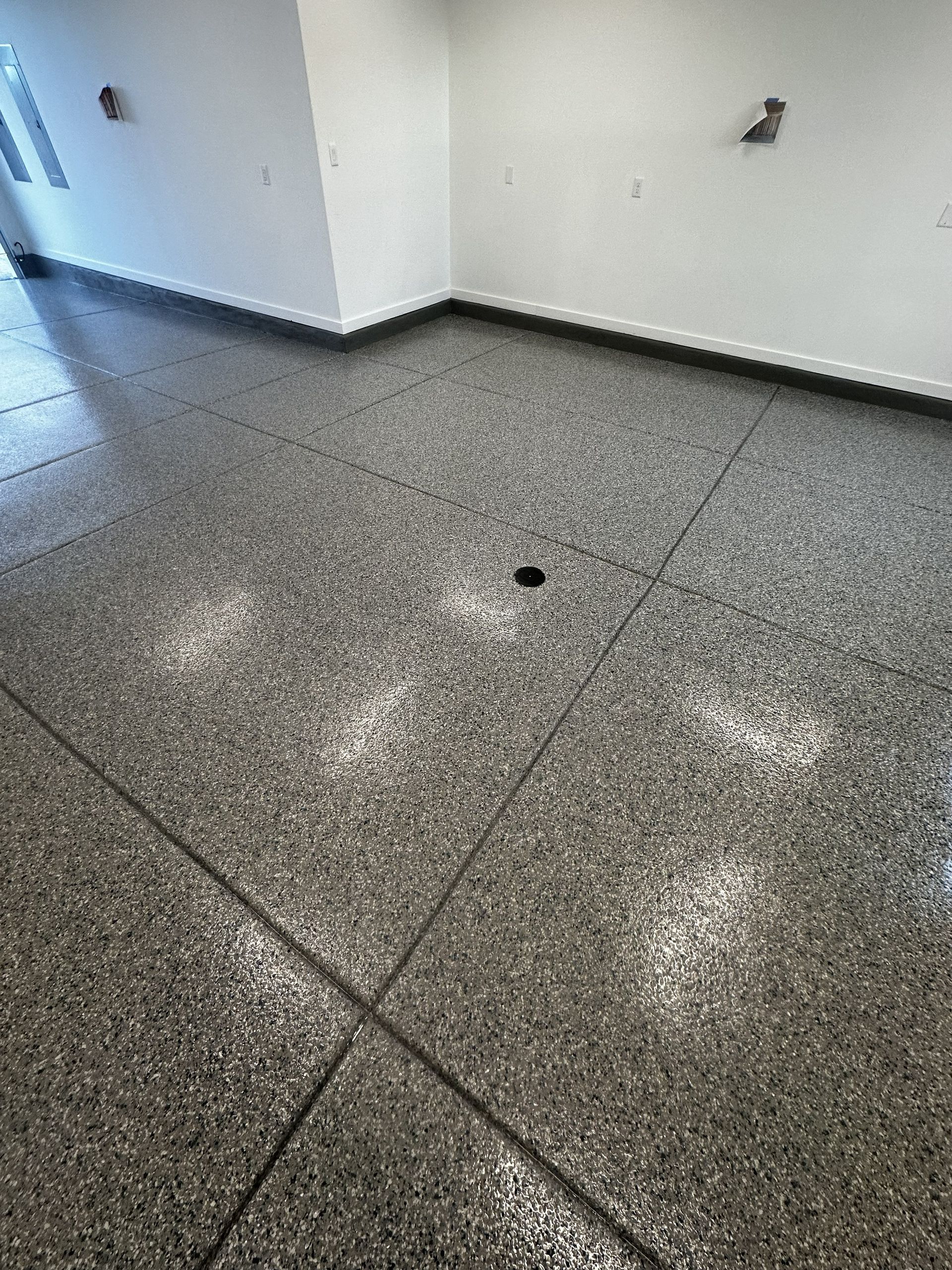Gray speckled concrete floor in a room, with diagonal lines and a drain in the center. White walls.