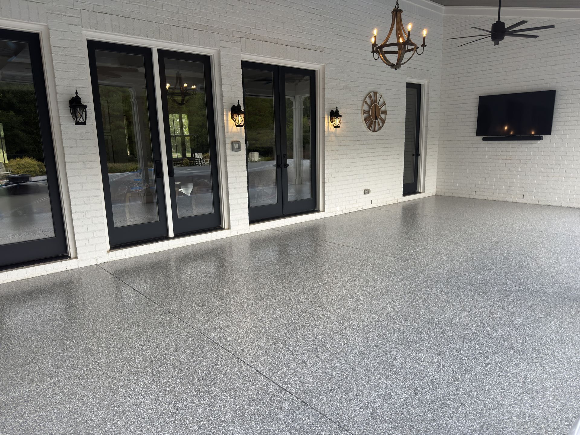 Spacious outdoor patio with shiny gray flooring, black framed doors, and white brick walls. Includes a chandelier and a mounted TV.