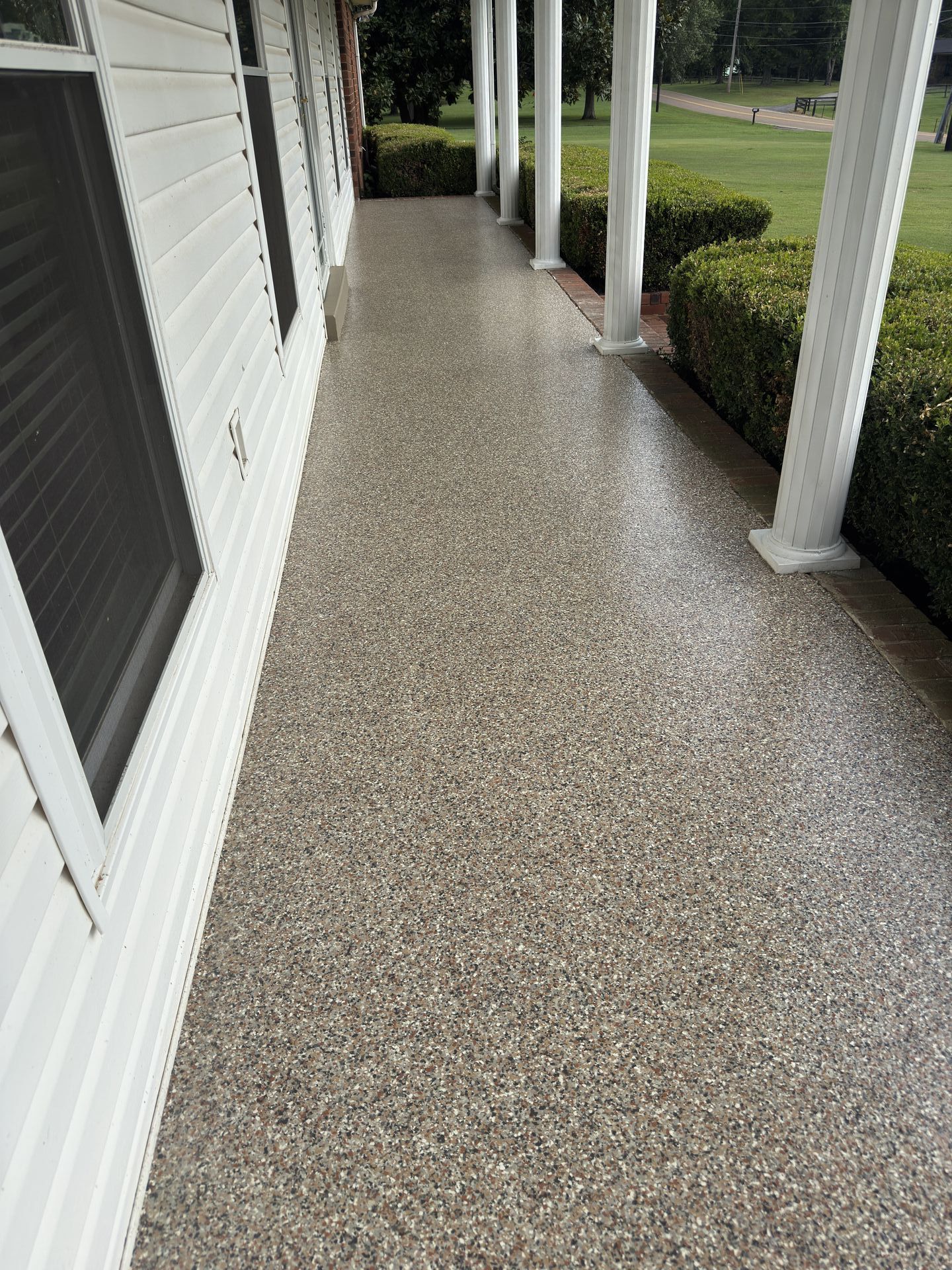 Covered porch with speckled, light gray concrete flooring. White siding and columns, green bushes.