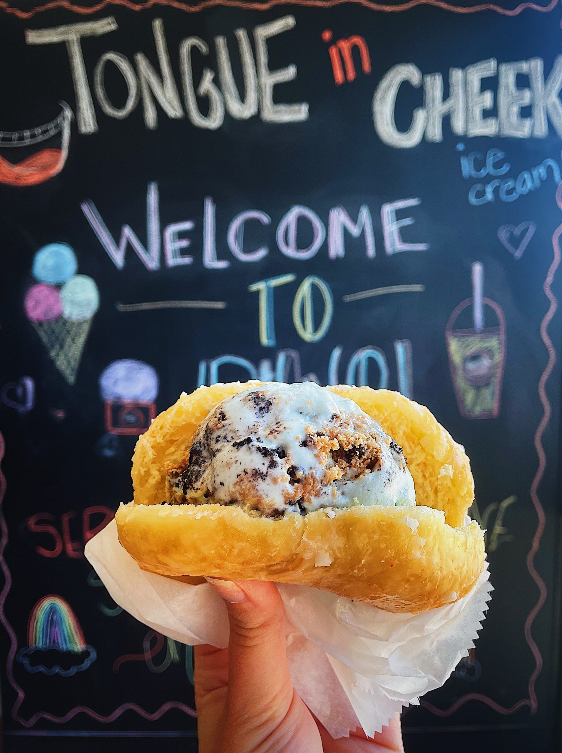 A person is holding a sandwich in front of a sign that says tongue in cheek