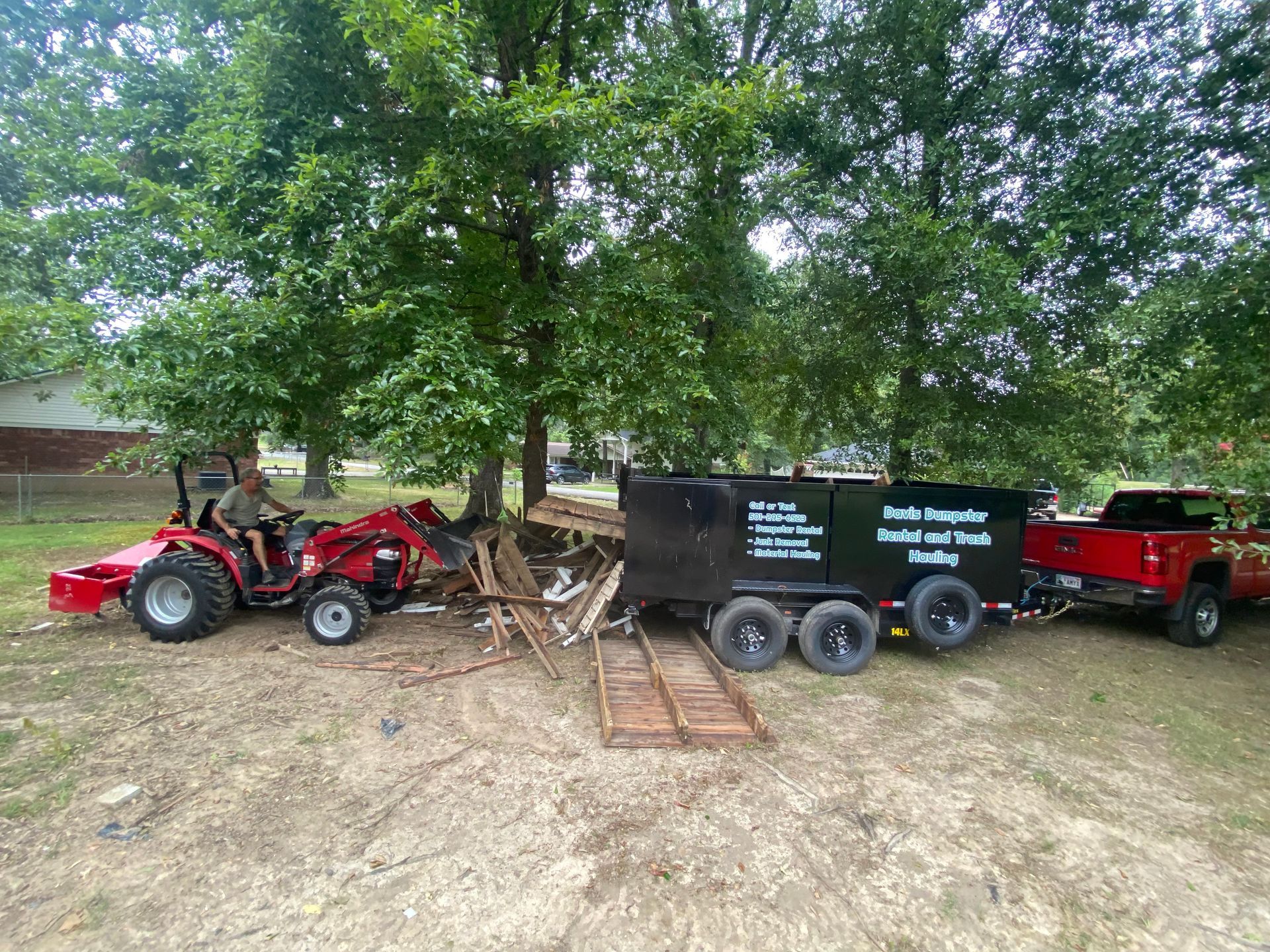 A red tractor pulling a trailer filled with debris, connected to a red truck, under trees.
