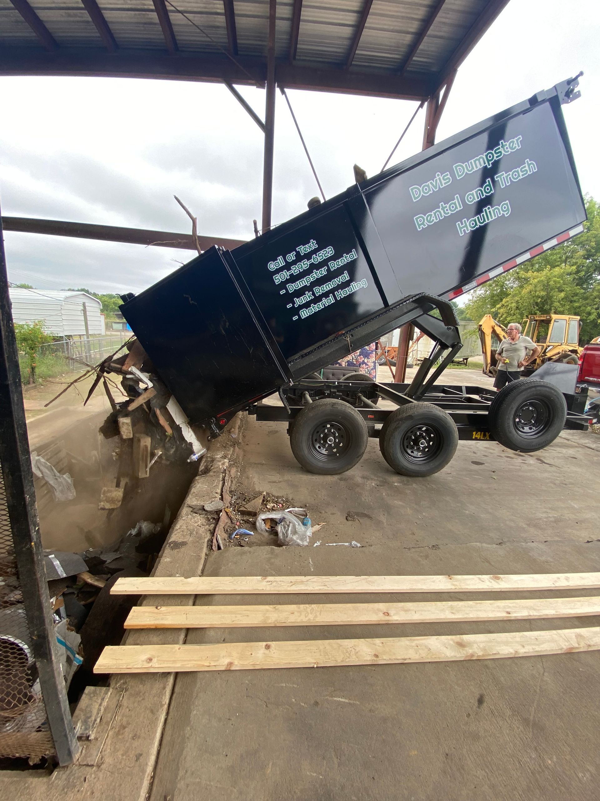 Black dump trailer unloading debris at a loading dock; gray building, brown planks.