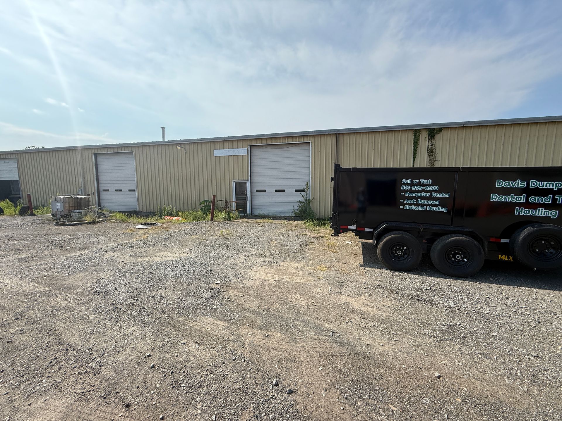 Exterior of a corrugated metal building with three garage doors. A black dump trailer is parked on gravel in front.