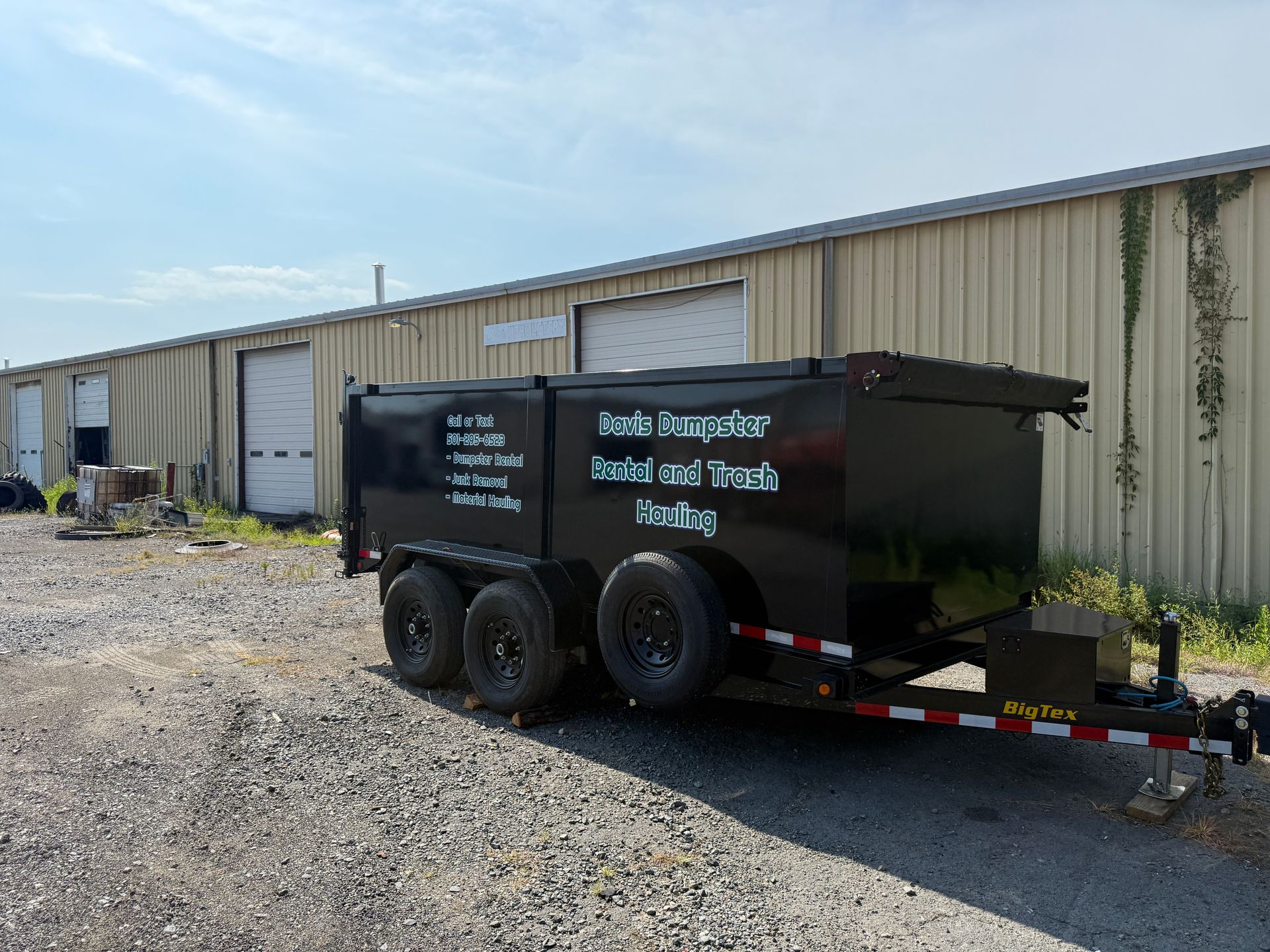 Black dumpster trailer parked in front of a metal building on a gravel lot under a blue sky.