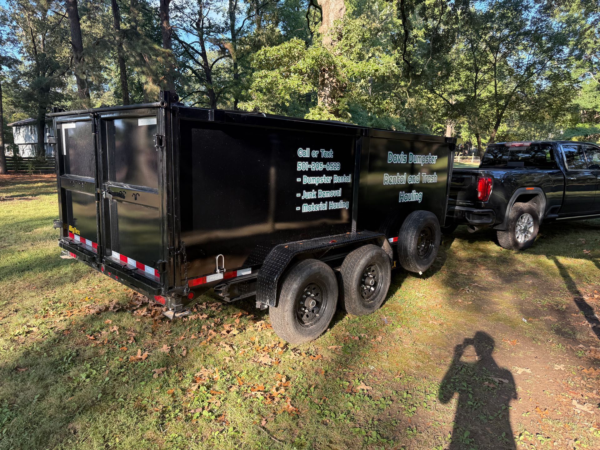 Black dump trailer hitched to a black pickup truck. Trailer has text advertising services.