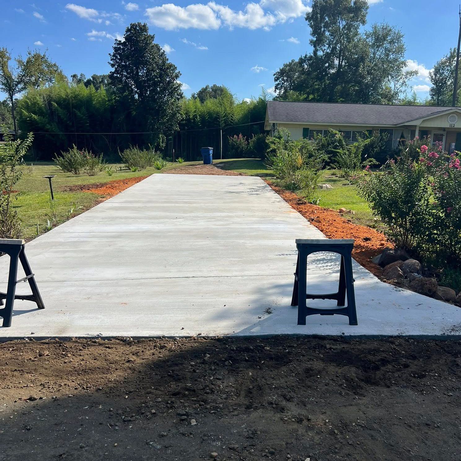 Newly poured concrete driveway leading to a yellow house with greenery under a blue sky.