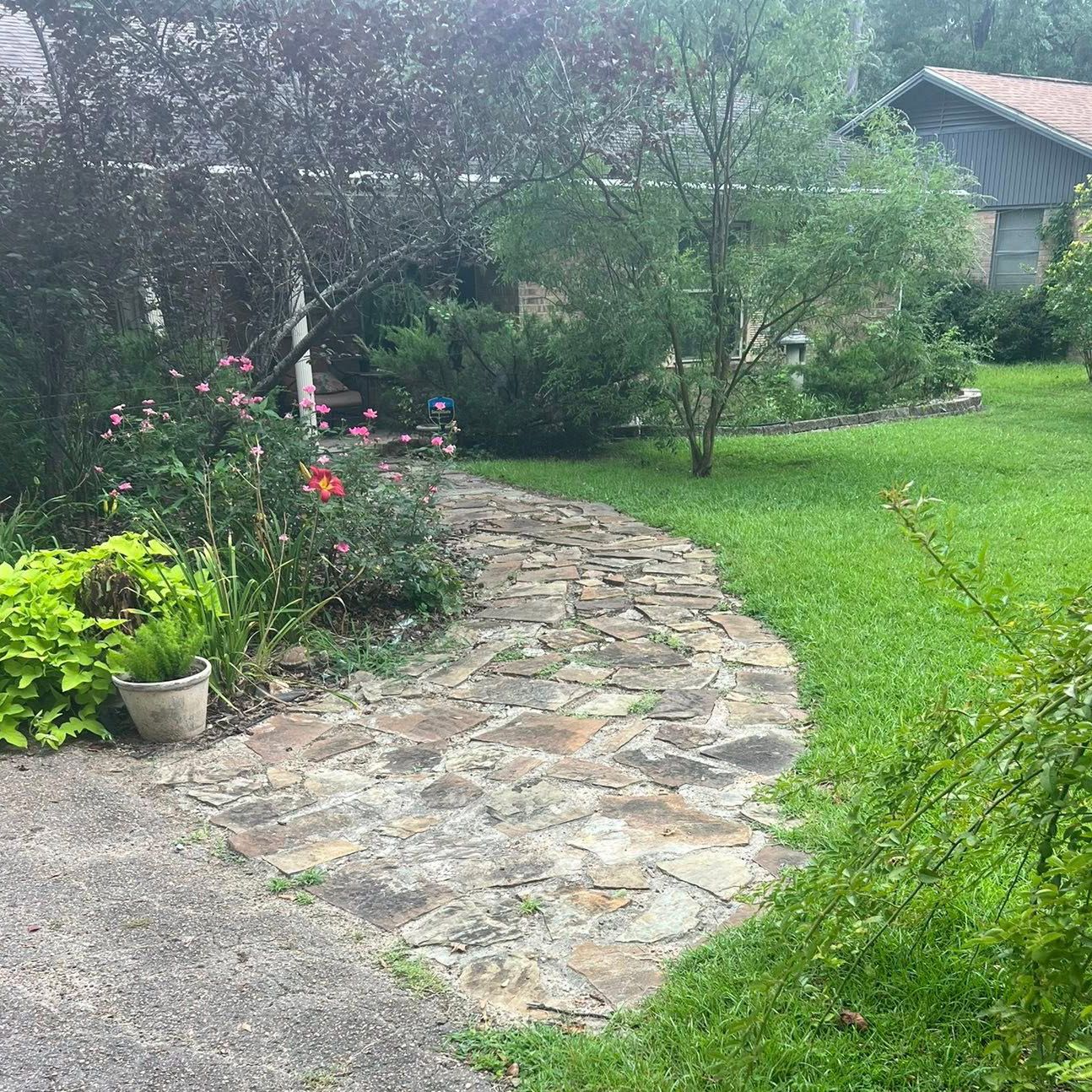 Stone path curves through a garden, flanked by green grass, flowers, and shrubs. Building in background.