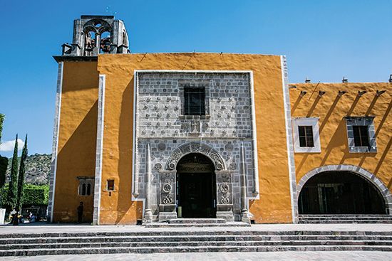 Fachada de iglesia de color amarillo mostaza con entrada de piedra ornamentada, puerta arqueada y torre contra un cielo azul.