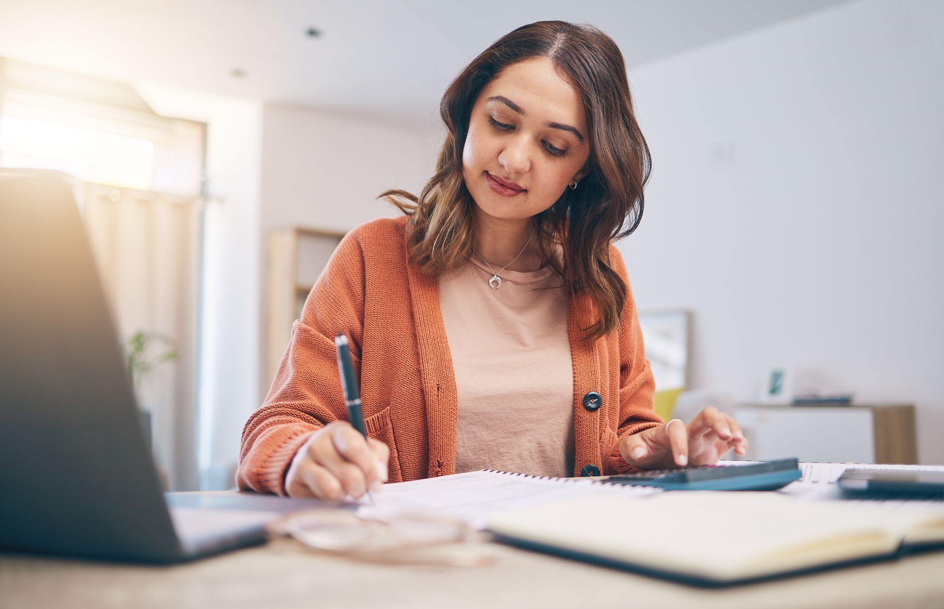 Woman calculating finances, writing notes, using a laptop, in a well-lit home setting.